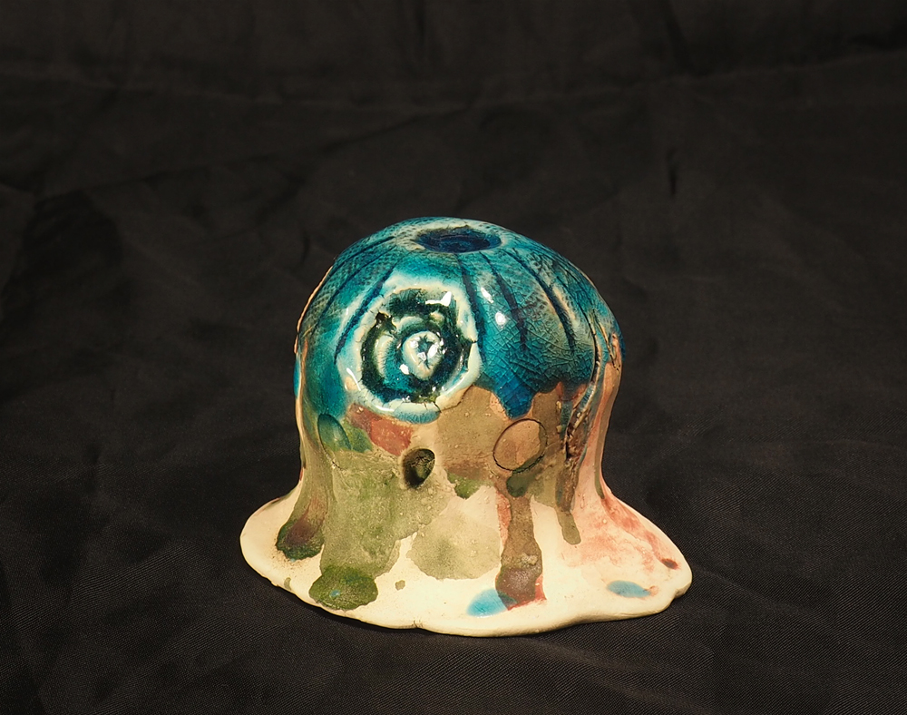 A small ceramic form that appears to be melting into the surface it sits on. The object is rounded like a baby doll's head and glazed in a drippy coating of bright blue, dusty green, and pale red.