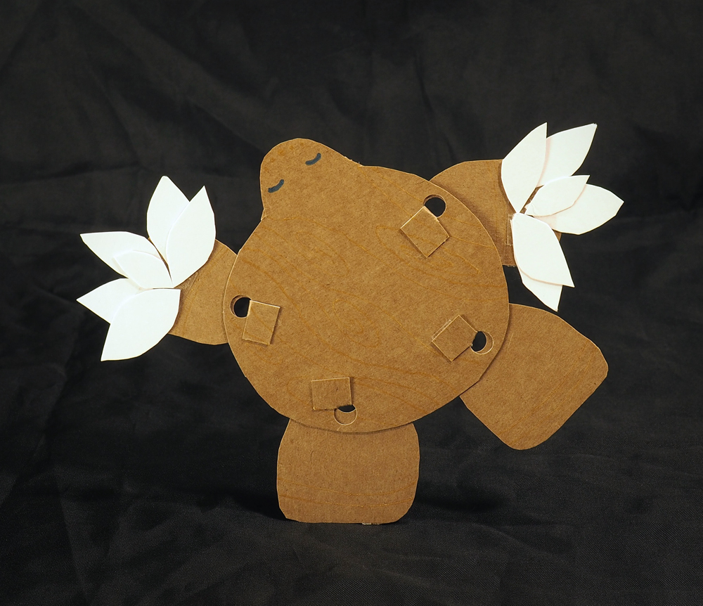 A thin, flat cardboard doll with a round shape and two closed eyes. Poseable legs and arms stick off of the brown body, and the arms end in clusters of pointed, white paper ovals, resembling flowers.