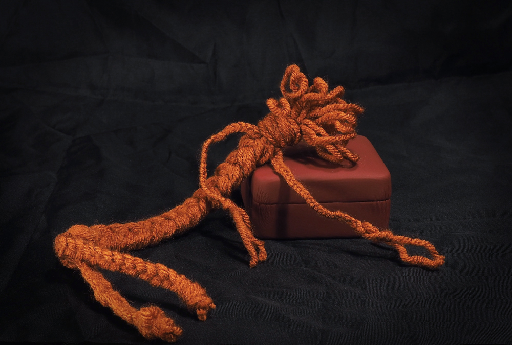 A braided yarn doll lounging dramatically against a small box. The doll is a dark pumpkin color, with a long, skinny body and limbs. The head is formed from a knot and several loops.