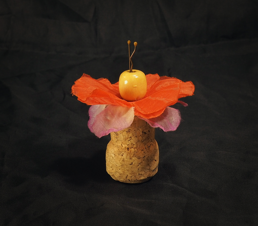 A tiny doll made from a champagne cork, red and purple fabric flowers, a round wood bead for a head, and a bobby pin sticking through the bead and flowers, into the top of the cork.