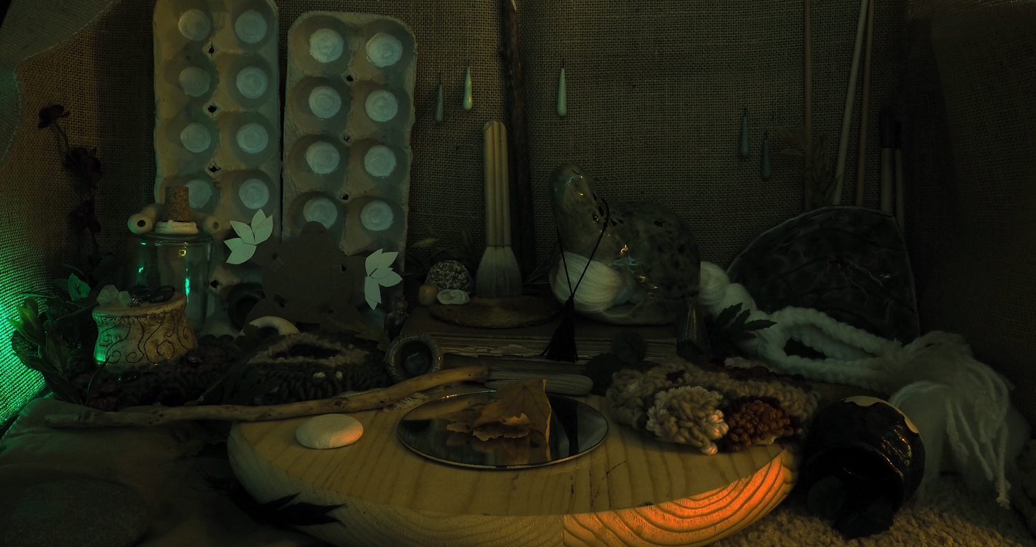 The Green Room with the lights turned off. A green glow shines through the burlap on the left, while a warm amber light reflects off of objects in the bottom right.