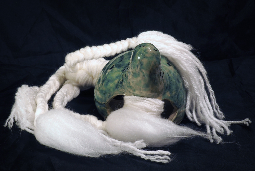 A creature-like doll with a ceramic body and a white braided yarn tail. The ceramic is glazed in a pale mottled green, and the long neck curves upward into a rounded head with two serene closed eyes. The tail is attached inside the body and flows outward, ending in four tufted ends.