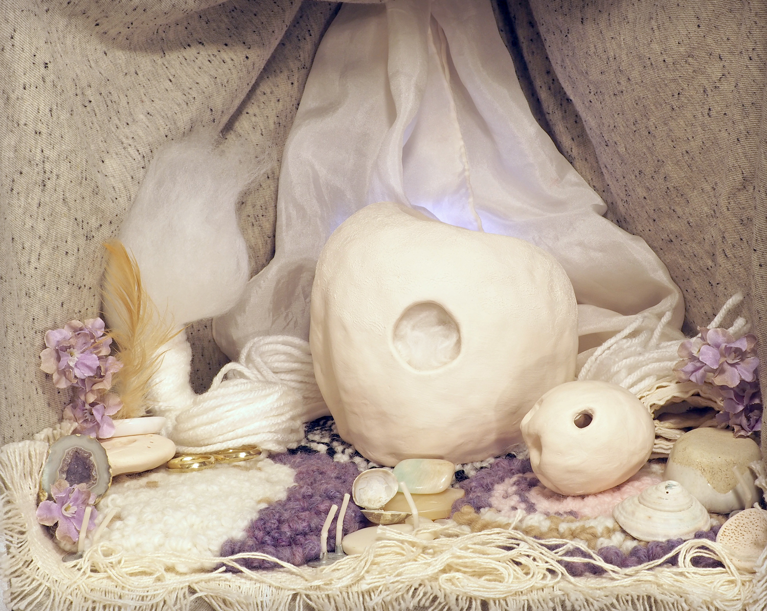 A room with a white and lavender color scheme. Speckled draped fabric makes up the cocoon-like walls, with light shining through a sheer white fabric at the back. The floor is covered with small yarn rugs. Purple flowers, white shells, feathers, and loose threads are scattered near the corners. The Wisp doll sits to one side, a puff of white fluff with a long tassel of yarn, it wraps around the back of the room. The Progenitor doll sits in the center, a large, round white ceramic form with a hole, through which more white fluff is visible. The Egg doll sits in front of it to the right, another ceramic form, egg-shaped and dented in on the front. The whole room looks soft and enveloping, with a delicate quality.