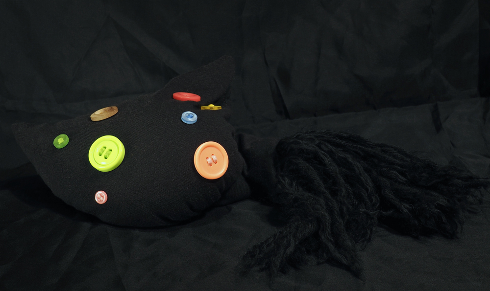 An amorphous black fabric doll that sits like a beanbag. Colorful buttons of various sizes are stitched onto one end, while the other end has many tufts of black yarn.