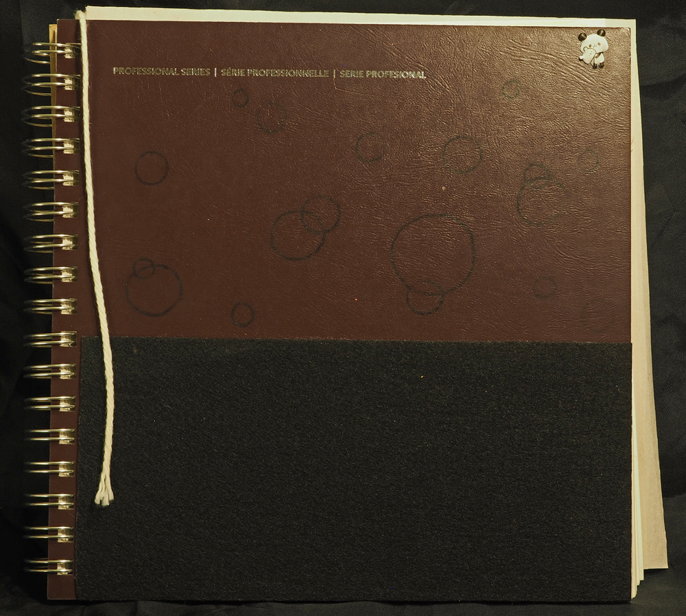 A spiral-bound sketchbook with a hard burgundy cover, the bottom half of which is covered in black felt. There are black circles drawn on the remaining space, a panda sticker in the top right corner, and a yarn bookmark.