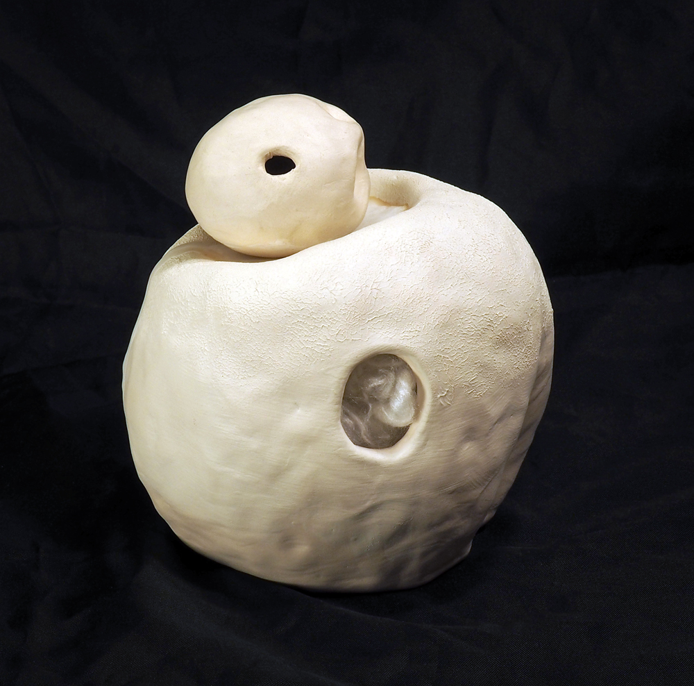 Two dolls, one nesting in the other. They are both plain white ceramic objects, with a rounded shape and a hole on either side. The smaller doll is similar in size and shape to an egg, with deep finger impressions on one end. The larger doll has white fiber fluff visible through its holes, an indent on the front, and a gentle dip on the top that is filled with wax, cushioning the smaller doll that sits in it.