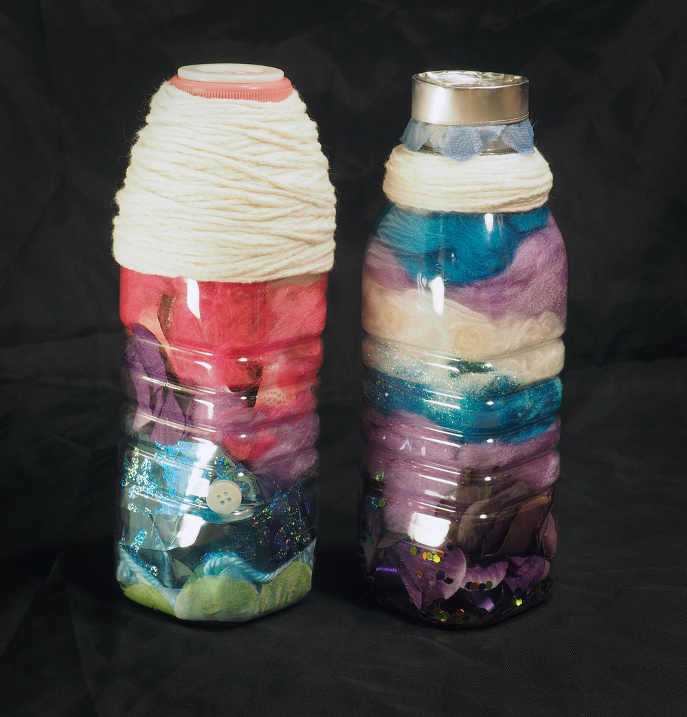 Two dolls standing side by side, each made from a plastic bottle. Both are relatively featureless and are filled with colorful objects like fake flowers, fiber fluff, glitter, and buttons. The doll on the left is thickly wrapped with white yarn at the top. The doll on the right is wrapped with a smaller amount of white yarn, with an aluminum cap and a blue fabric flower on top.