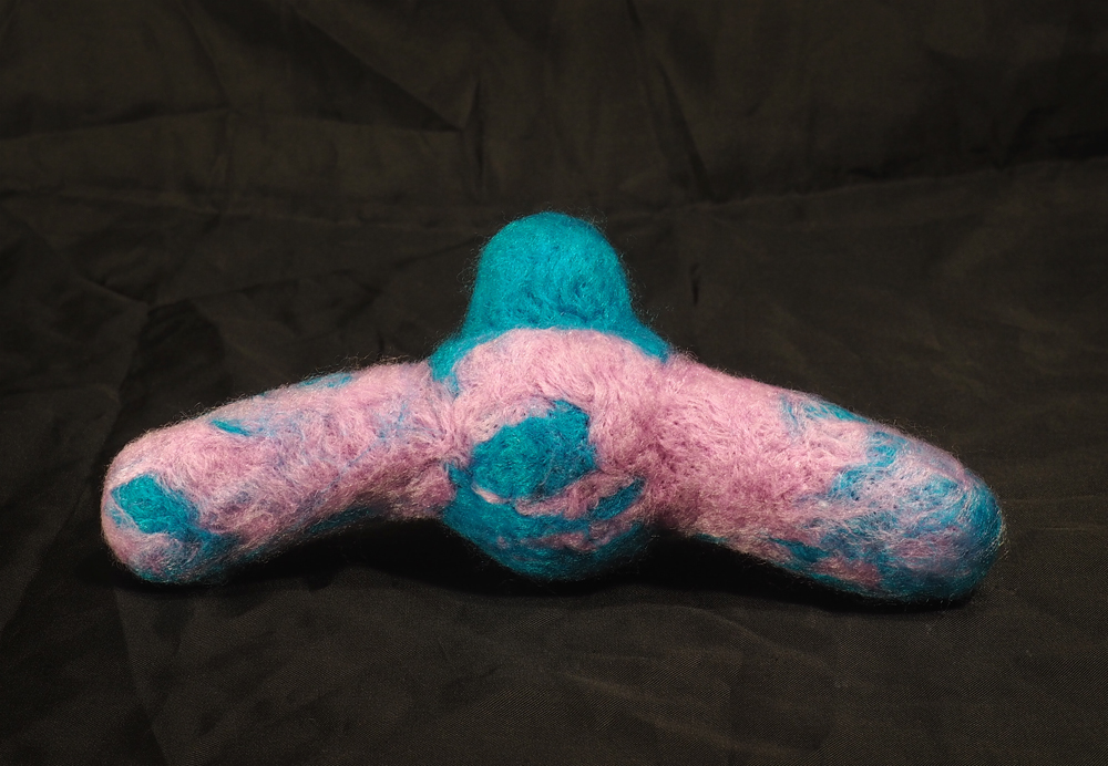 A blue and purple needle felted doll with a round body, small featureless head, and two thick limbs sticking off the sides. The limbs are rounded at the tips.